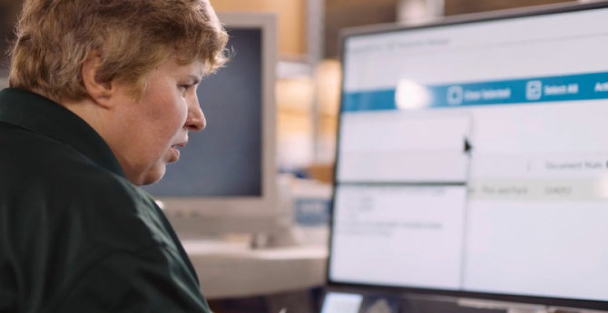 A worker in a green shirt focuses on a computer screen displaying software menus, with a second monitor in the background, in an office environment.