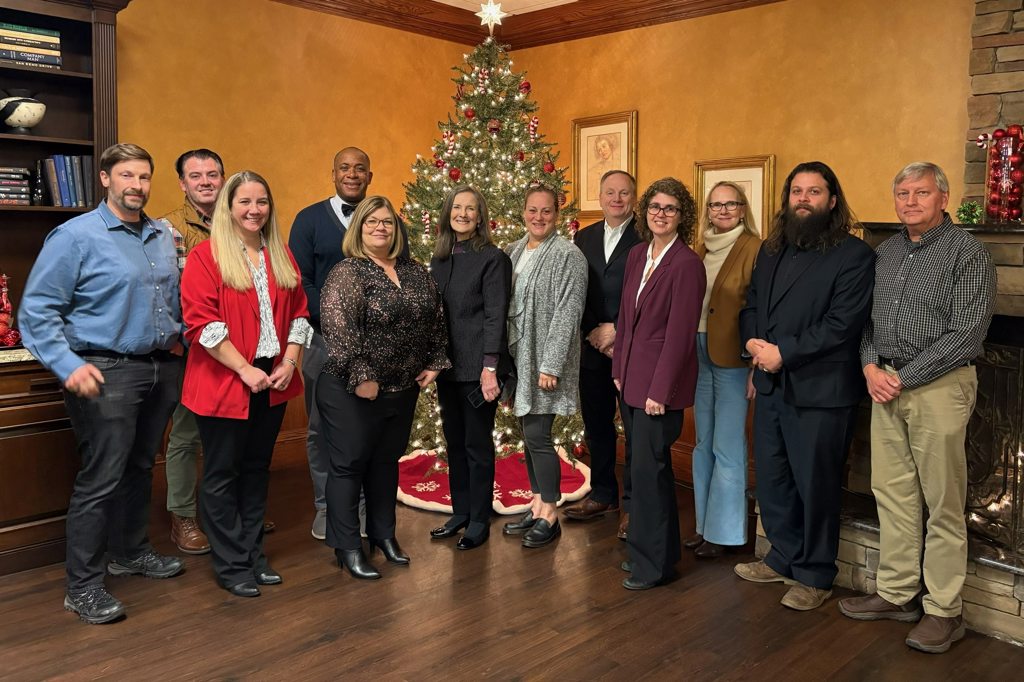 twelve smiling professionals (men and women) posing in warm-lit room with decorated Christmas tree; casual business attire, wooden floor.