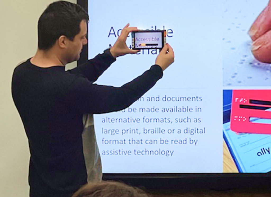 Presenter holding accessible tablet; slide on providing materials in large print, braille, or digital formats readable by assistive technology.