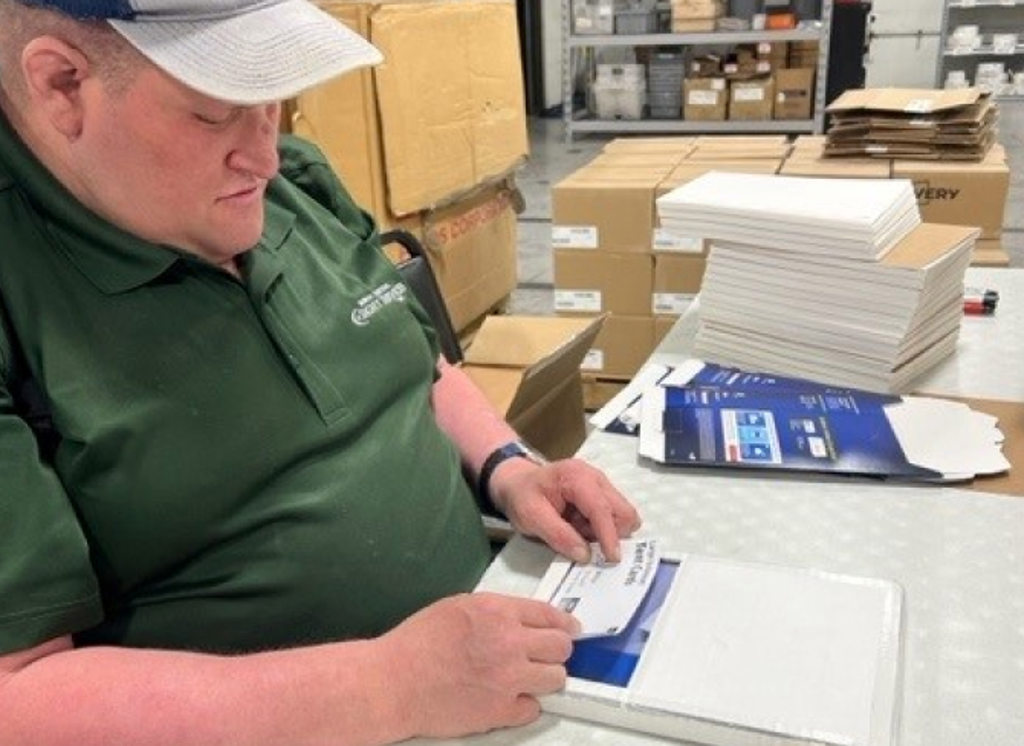 A worker in a green shirt and cap packaging items at a table in a warehouse, surrounded by boxes and supplies, focusing on the task.