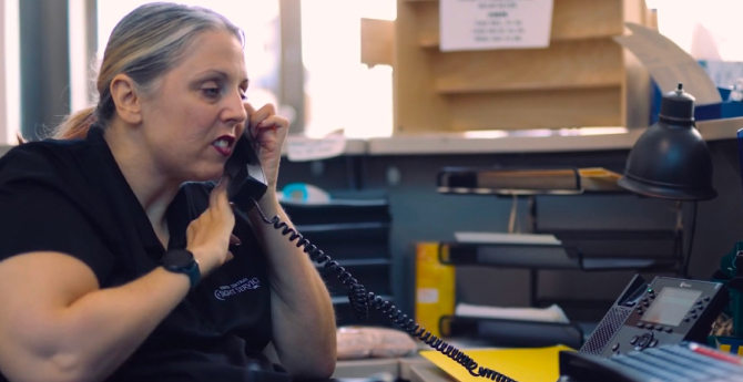 A woman in a black uniform with a logo speaks on a corded phone in an office, surrounded by desk supplies and equipment, showing focus at work.
