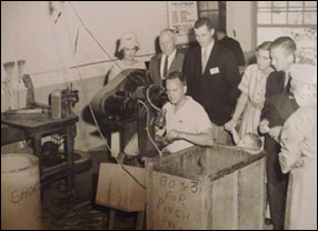 a black and white photo of a blind employee working on the industry line with people standing behind him observing. 